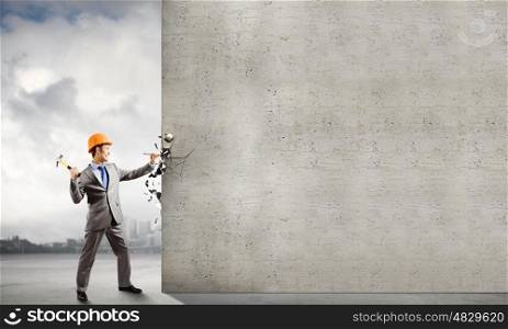 Man with hammer. Businessman in helmet hammering nail in brick wall