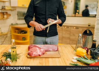 Man with knife prepares to cut raw meat, kitchen interior on background. Chef cooking tenderloin with vegetables, spices and herbs. Man with knife prepares to cut raw meat
