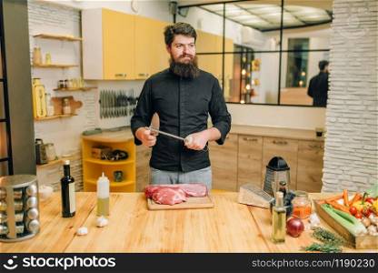 Man with knife prepares to cut raw meat, kitchen interior on background. Chef cooking tenderloin with vegetables, spices and herbs