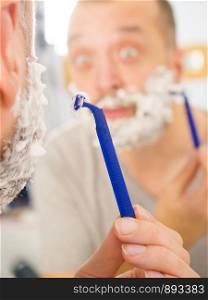 Man with lots of shaving foam on his face standing in bathroom, looking at mirror, shaving his face beard. Skincare. Guy shaving his beard in bathroom