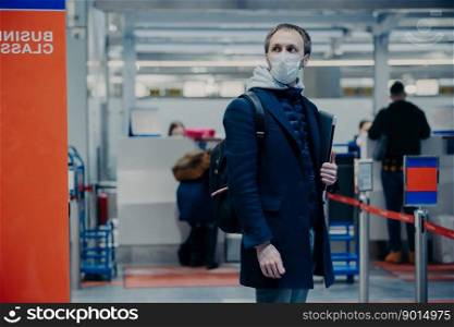 Man with luggage, wears medical protection mask, poses in airport terminal, travels by airplane during coronavirus outbreak, prevents disease, ready for fight and health checkpoint. Evacuated tourist