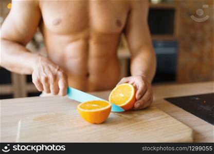 Man with naked body cuts orange on the kitchen. Nude male person preparing breakfast at home, food preparation without clothes