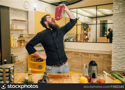Man with opened mouth holds piece of fresh raw meat, kitchen interior on background. Chef cooking tenderloin with vegetables, spices and herbs