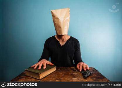 Man with paper bag over his head sitting at table with book and revolver