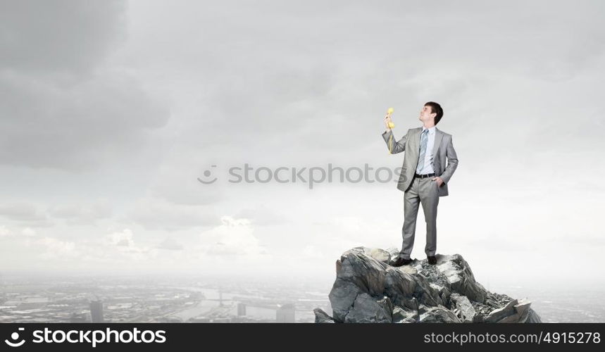 Man with phone receiver. Smiling businessman talking on yellow phone handset on rock top