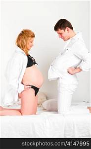 man with pillow under shirt and his pregnant wife are comparing their bellies.