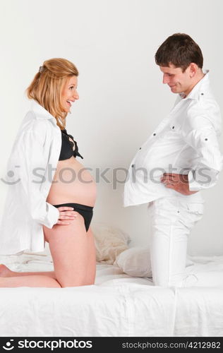 man with pillow under shirt and his pregnant wife are comparing their bellies.