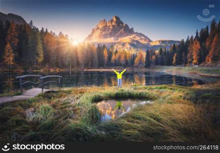 Man with raised up arms on Antorno lake with reflection of Tre Cime Di Lavaredo at sunrise in autumn in Dolomites, Italy. Landscape with happy man, green grass, forest, pond, sun and high rocks