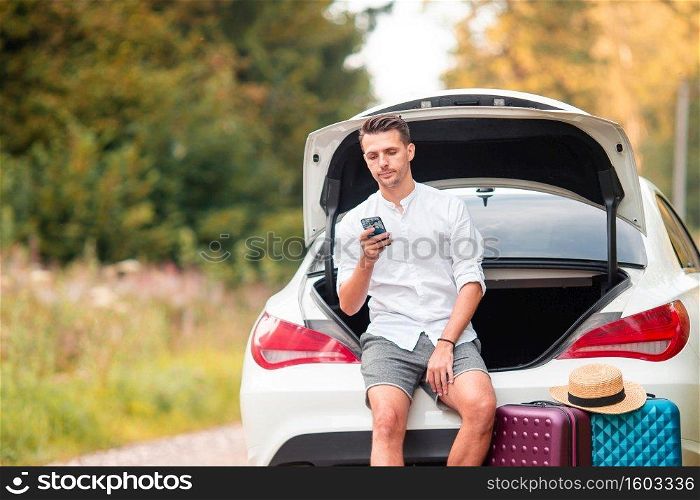 Man with smartphone sits in trunk of his car during his weekend. Young couple tourist enjoying on summer vacation