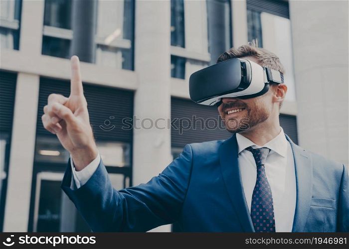 Man with stubble dressed in blue formal suit, wearing VR glasses, viewing something in virtual reality, pointing up with his finger and using it to control, standing next to building. Smiling bearded businessman with stubble wearing VR glasses, viewing something in virtual reality