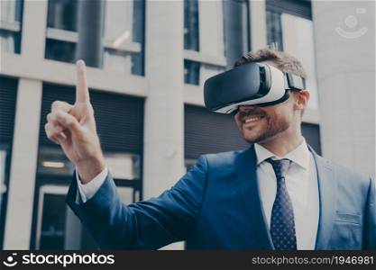 Man with stubble dressed in blue formal suit, wearing VR glasses, viewing something in virtual reality, pointing up with his finger and using it to control, standing next to building. Smiling bearded businessman with stubble wearing VR glasses, viewing something in virtual reality