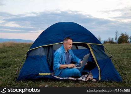 Man working on laptop in tent in nature. Young freelancer sitting in camp. outdoor activity in summer. internet 5G. Man working on laptop in tent in nature. Young freelancer sitting in camp. Relaxing in mountains. Remote work, outdoor activity in summer. internet 5G.