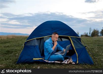 Man working on laptop in tent in nature. Young freelancer sitting in camp. Relaxing in mountains. Remote work, outdoor activity in summer. Happy male work on vacation. Man working on laptop in tent in nature. Young freelancer sitting in camp. Relaxing in mountains. Remote work, outdoor activity in summer. Happy male work on vacation.