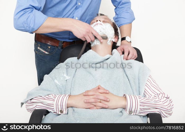 Man wrapped in towel being shaved with cut throat razor by barber over colored background