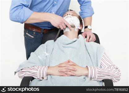 Man wrapped in towel being shaved with cut throat razor by barber over colored background