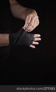 man wraps his hands in black textile bandage for sports, black background