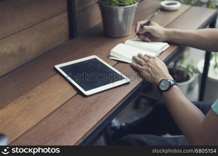 man writing on notebook with digital tablet on wooden table, selective focus and vintage tone