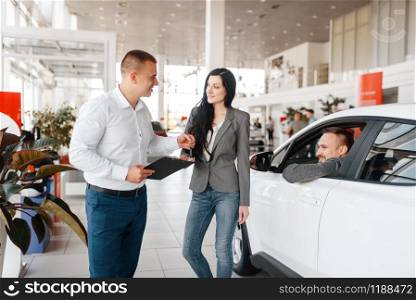 Manager and happy couple near the new car in showroom. Male and female customers looks vehicle in dealership, automobile sale, auto purchase