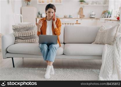 Manager explains to customer on phone. Young european woman in orange shirt is sitting on couch at home holding computer. Employee is talking to boss on telephone. Remote work on quarantine.. Manager explains to customer on phone. Young european woman works at home on quarantine.