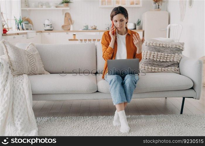 Manager explains to customer on phone. Young european woman in orange shirt is sitting on couch at home holding computer. Employee is talking to boss on telephone. Remote work on quarantine.. Manager explains to customer on phone. Young european woman works at home on quarantine.