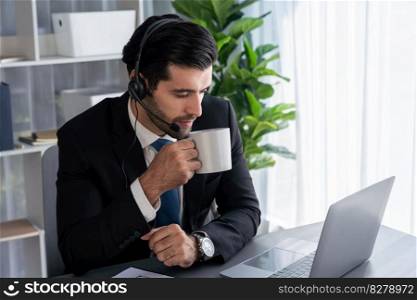 Manager of call center operator office sitting on his desk with his coffee while working on laptop. Office worker wearing headset and black suit working on customer support or telemarketing. fervent. Manager of call center operator office sitting on his desk with coffee. fervent