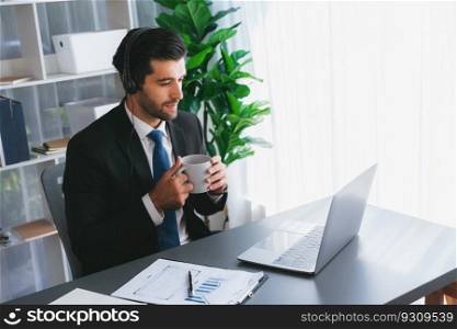 Manager of call center operator office sitting on his desk with his coffee while working on laptop. Office worker wearing headset and black suit working on customer support or telemarketing. fervent. Manager of call center operator office sitting on his desk with coffee. fervent