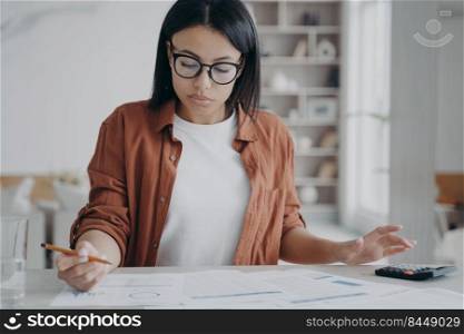 Manager or business assistant is sitting at the desk and doing her job with paperwork. Concentrated girl, businesswoman is examining reports. Young spanish woman in glasses working remote from home.. Manager or business assistant is sitting at the desk and doing her job with paperwork at home.