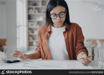 Manager or business assistant is sitting at the desk and doing her job with paperwork. Concentrated girl, businesswoman is examining reports. Young spanish woman in glasses working remote from home.. Manager or business assistant is sitting at the desk and doing her job with paperwork at home.