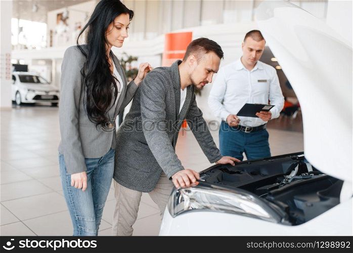 Manager shows the couple a new car in showroom. Male and female customers looks vehicle in dealership, automobile sale, auto purchase. Manager shows the couple a new car in showroom