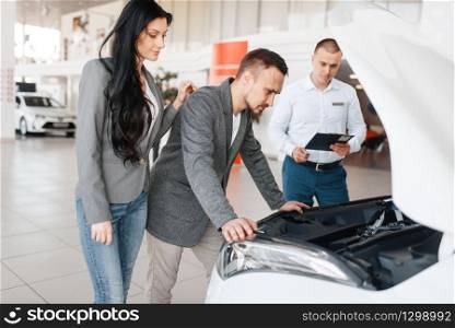 Manager shows the couple a new car in showroom. Male and female customers looks vehicle in dealership, automobile sale, auto purchase. Manager shows the couple a new car in showroom