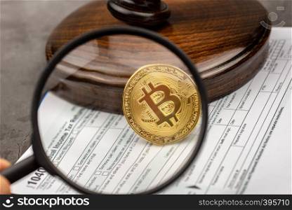 Mand hand holds magnifying glass with view of Bitcoin cryptocurrency in ront of gavel.Tax paying concept.