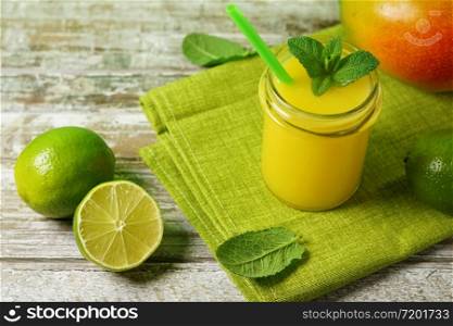 Mango lime smoothie refreshing tropical drink front top view of a glass jar with mint leaves, mango and sliced lime fruits on a wooden rustic table.