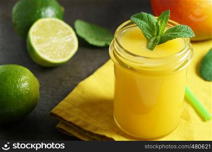 Mango lime smoothie refreshing tropical drink front view close up of a glass jar with mint leaves and mango and sliced lime fruits on background.