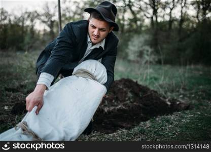 Maniac with victim&rsquo;s body wrapped in a canvas against a grave, serial murderer concept, crime horror