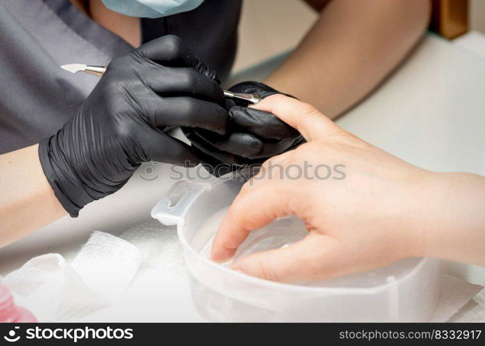 Manicurist removing cuticle from female nails by metal pusher during soaking fingernails in the bath at nail salon. Manicurist removing cuticle from nails
