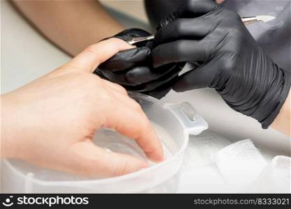 Manicurist removing cuticle from female nails by metal pusher during soaking fingernails in the bath at nail salon. Manicurist removing cuticle from nails