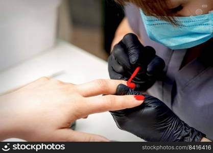 Manicurist with protective mask and gloves applying red nail polish to female nails in nail salon. Manicurist applying red nail polish