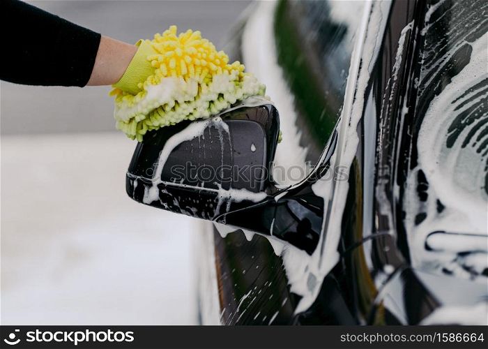 Mans hand holding sponge for washing car. Black auto with soap bubbles. Cleaning concept. Worker cleaning automobile