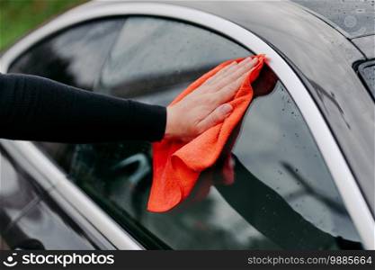 Mans hand with rag cleaning side window of black car. Professional auto wash concept. Regular wash up. Wiping water droplets on vehicle