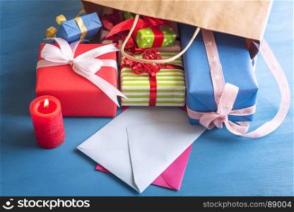 Many and colorful presents spilled out of a bag on a blue wooden table, falling over empty envelopes, near a lit red candle.