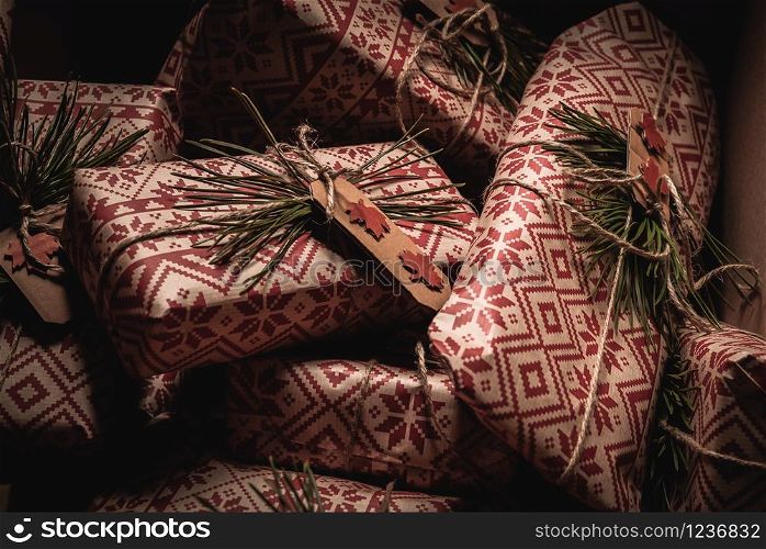 Many christmas presents piled up in a box with creative handmade decorative rustic diy gift wrapped in red retro wrapping paper with natural vintage twine and spruce twigs as decor, hidden away until Xmas Eve.