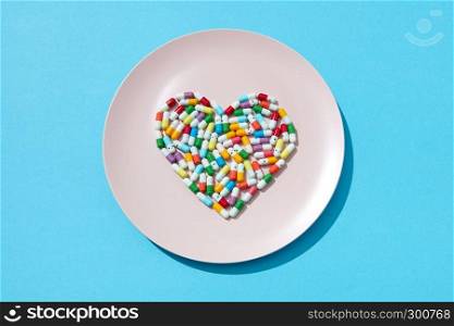 Many different pills and supplements in a shape of a heart on a white plate on a blue background. Cardiovascular disease prevention. Flat lay. Heart made from medicine pills and capsules on a white plate on a blue background, copy space. Colorful food supplement pills. Top view.