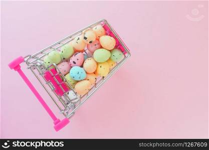 many multi-colored miniature artificial eggs in a miniature food cart on a pink background. View from above. Easter sale concept.