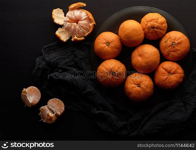 many orange ripe round whole tangerines in a plate on a black wooden table, ripe and juicy fruits, top view