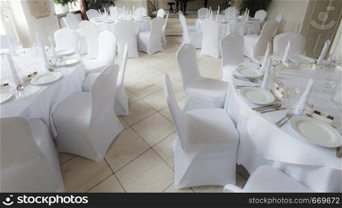Many wedding white chairs and round tables in small light ballroom. Bridal place decorations.. Many wedding chairs and tables