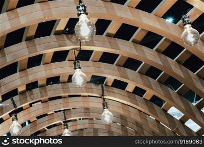 Many White Light Bulbs are hung on the Ceiling of the Wooden Curved Pergola Structure. Light Bulbs in Wooden Facade Architecture. Space for text, Selective focus.
