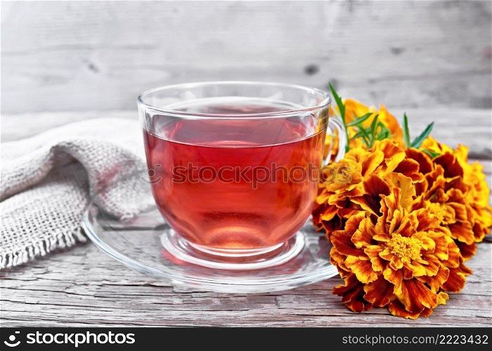 Marigold herbal tea in a glass cup and saucer, fresh flowers, burlap on wooden board background