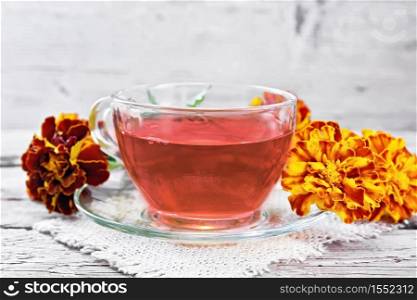 Marigold herbal tea in a glass cup and saucer on a napkin of sackcloth, fresh flowers on wooden board background