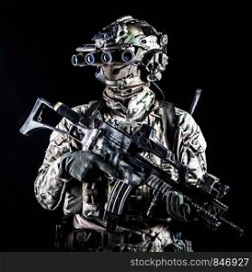 Marine rider in camouflage uniform and face mask, patrolling in darkness with quad-tube night vision goggles on battle helmet, holding modern assault rifle, low key studio portrait on black background. Marine rider with rifle and night vision goggles