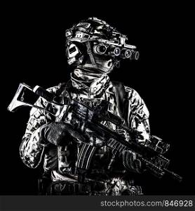 Marine rider in camouflage uniform and face mask, patrolling in darkness with quad-tube night vision goggles on battle helmet, holding modern assault rifle, low key studio portrait on black background. Marine rider with rifle and night vision goggles